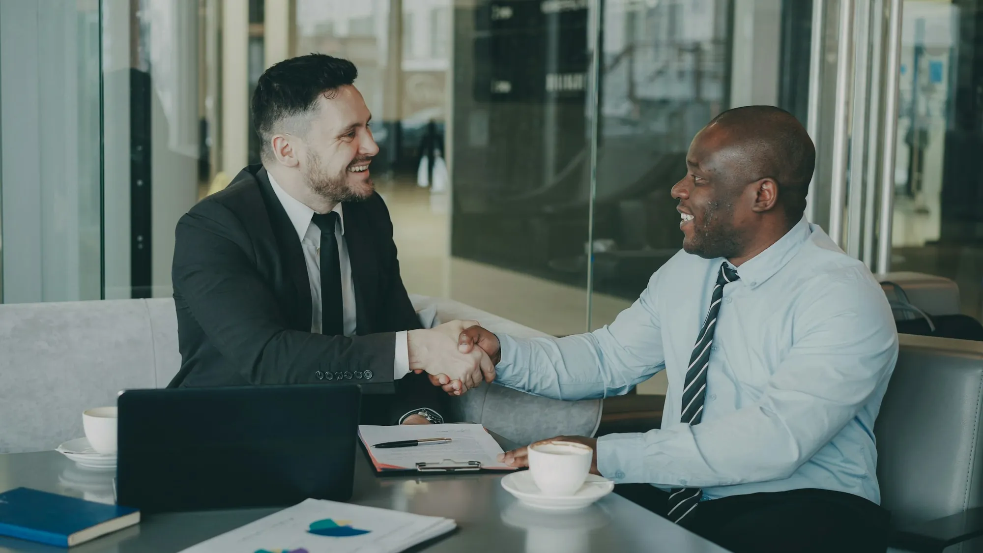 Two smiling men in suits shaking hands at a meeting table